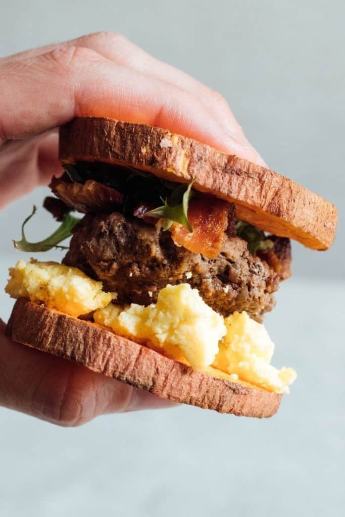 A hand holds a breakfast sandwich including sausage, eggs, bacon, and lettuce on a sweet potato bun.