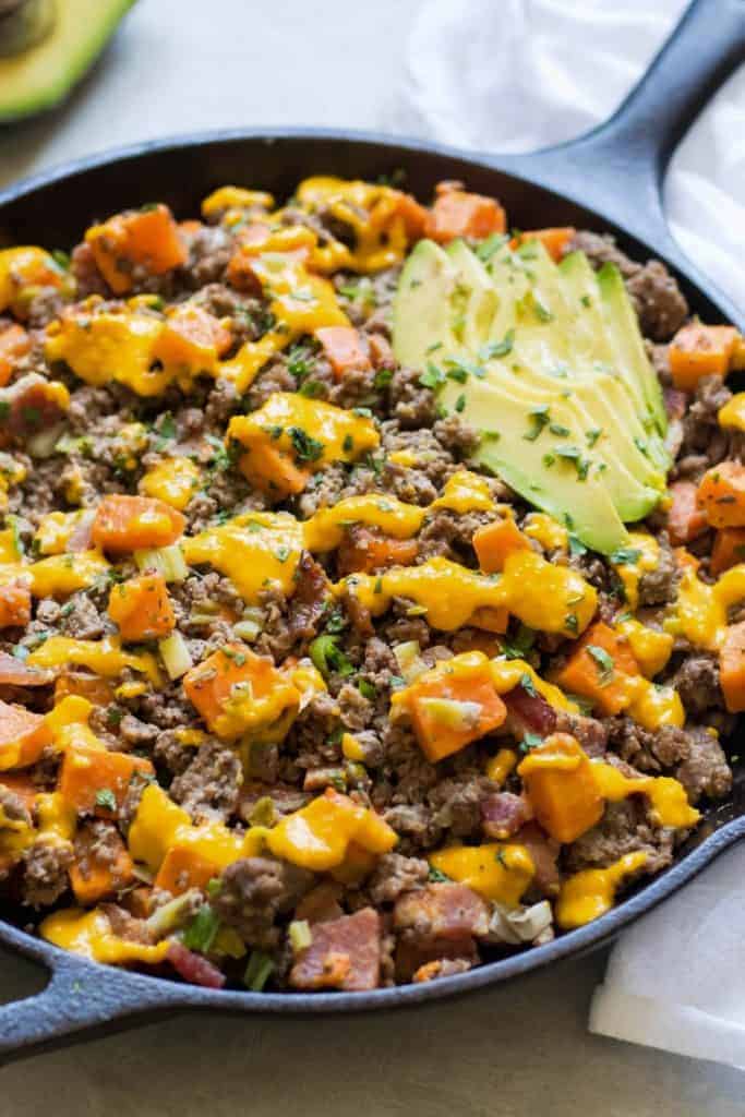 a skillet of ground beef, bacon, dairy free cheese sauce, and avocado sprinkled with cilantro on a grey background comes together to make a Bacon Cheeseburger Casserole