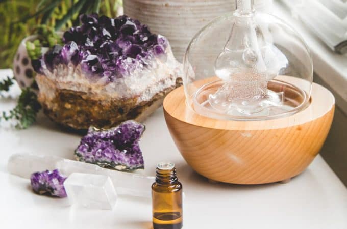A glass diffuser and essential oil bottle on a white table with a amethyst geode crystal.
