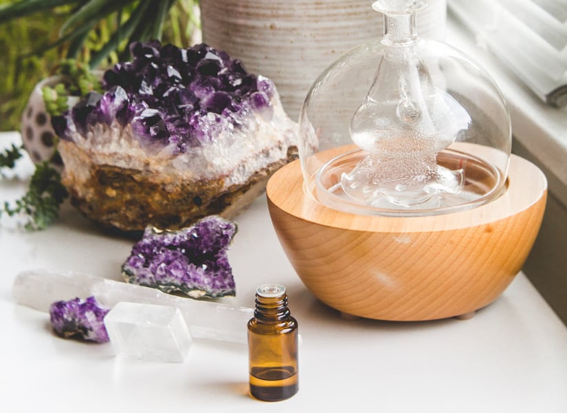 A glass diffuser and essential oil bottle on a white table with a amethyst geode crystal.
