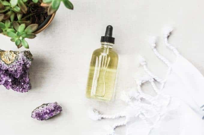 Light gray table with a bottle of face oil for the oil cleansing method, along with an amethyst geode, a turkish towel, and plant.