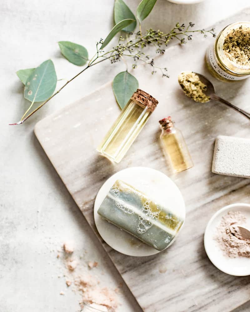 Bottle of face oil for the oil cleansing method on a light gray table with a face mask, eucalyptus, and a bar of soap.
