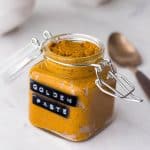Glass jar full of turmeric paste on a white counter with a golden spoon next to it.