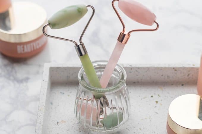 A jade and rose quartz roller in a glass cup on a terrazzo tray and marble countertop with pink skincare products from the Beautycounter Countertime line.