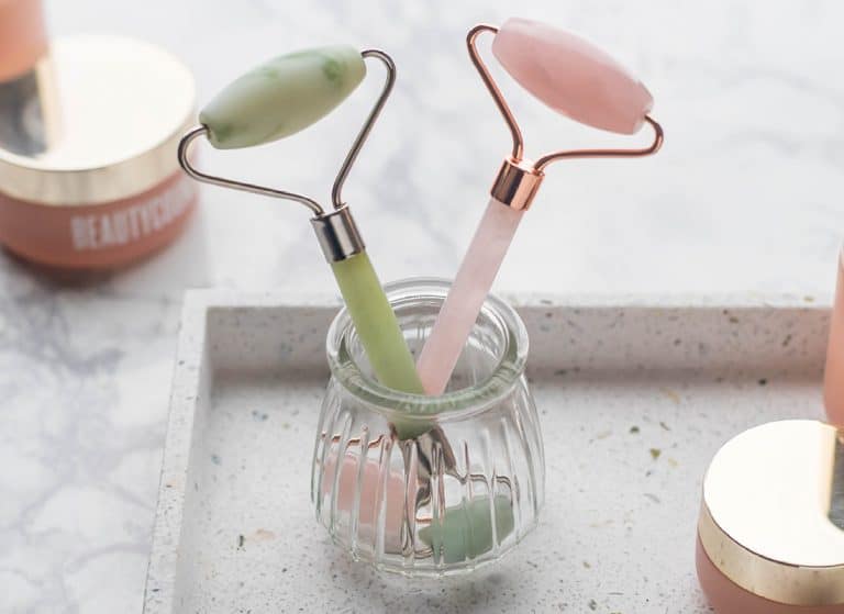 A jade and rose quartz roller in a glass cup on a terrazzo tray and marble countertop with pink skincare products from the Beautycounter Countertime line.
