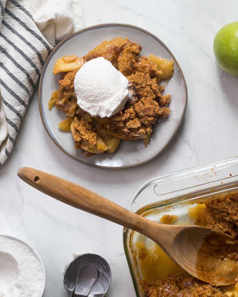 A marble table with a plate of Paleo Apple Crisp and dairy free coconut milk ice cream on top. There is a striped napkin, and off to the side you see the baking dish of apple crisp, an ice cream scoop, and a green apple.