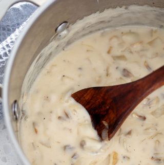 A metal stock pot with cream of mushroom soup, a blue towel, and a wooden spoon on a white countertop.