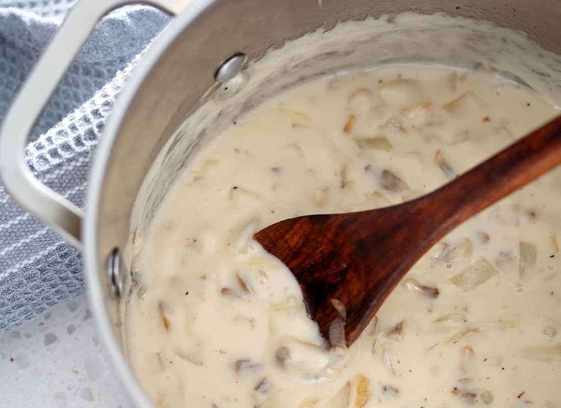 A metal stock pot with cream of mushroom soup, a blue towel, and a wooden spoon on a white countertop.