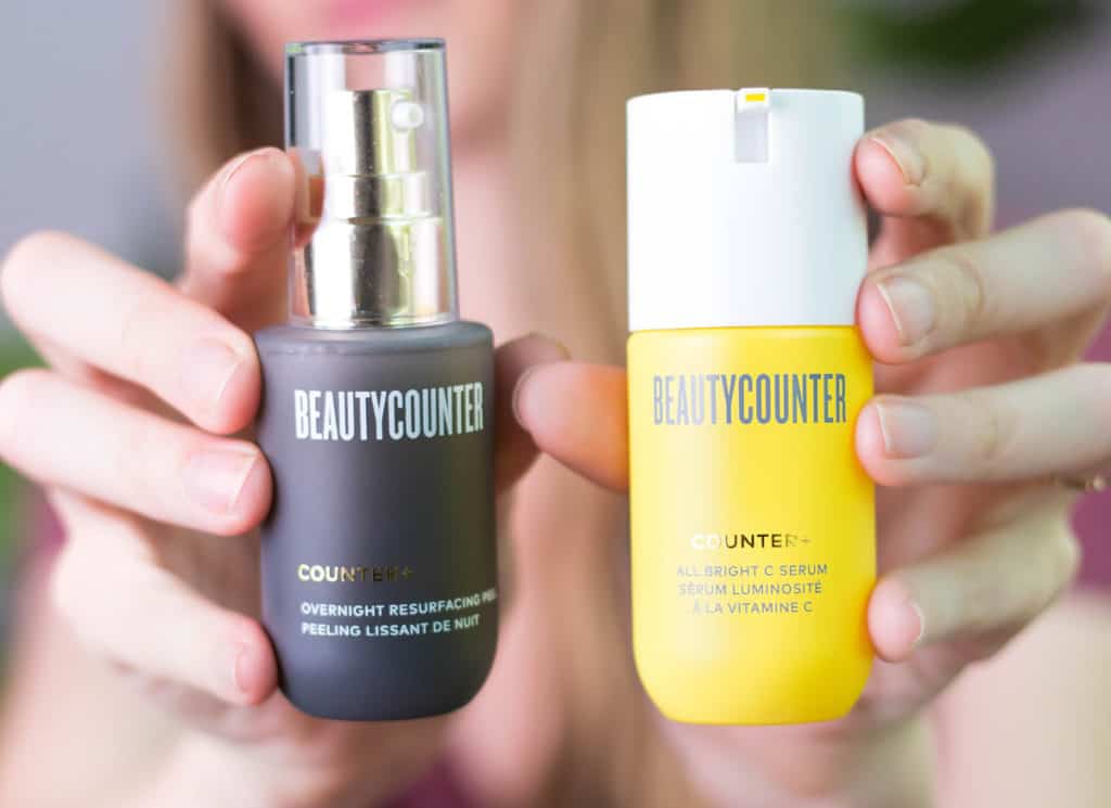 A woman in a pink shirt holds a jar of Beautycounter's All Bright C Serum and a jar of the Overnight Resurfacing Peel. 